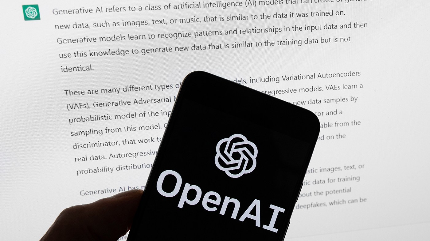 The OpenAI logo is displayed on a mobile phone in front of a computer screen with output from ChatGPT, March 21, 2023, in Boston.