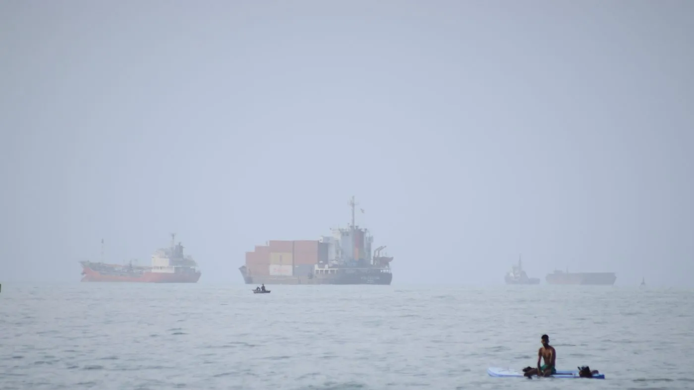 A man sits on a surfboard as a container ship and other commercial vessels appear anchored in the Strait of Hormuz off Bandar Abbas, Iran, July 27, 2026.