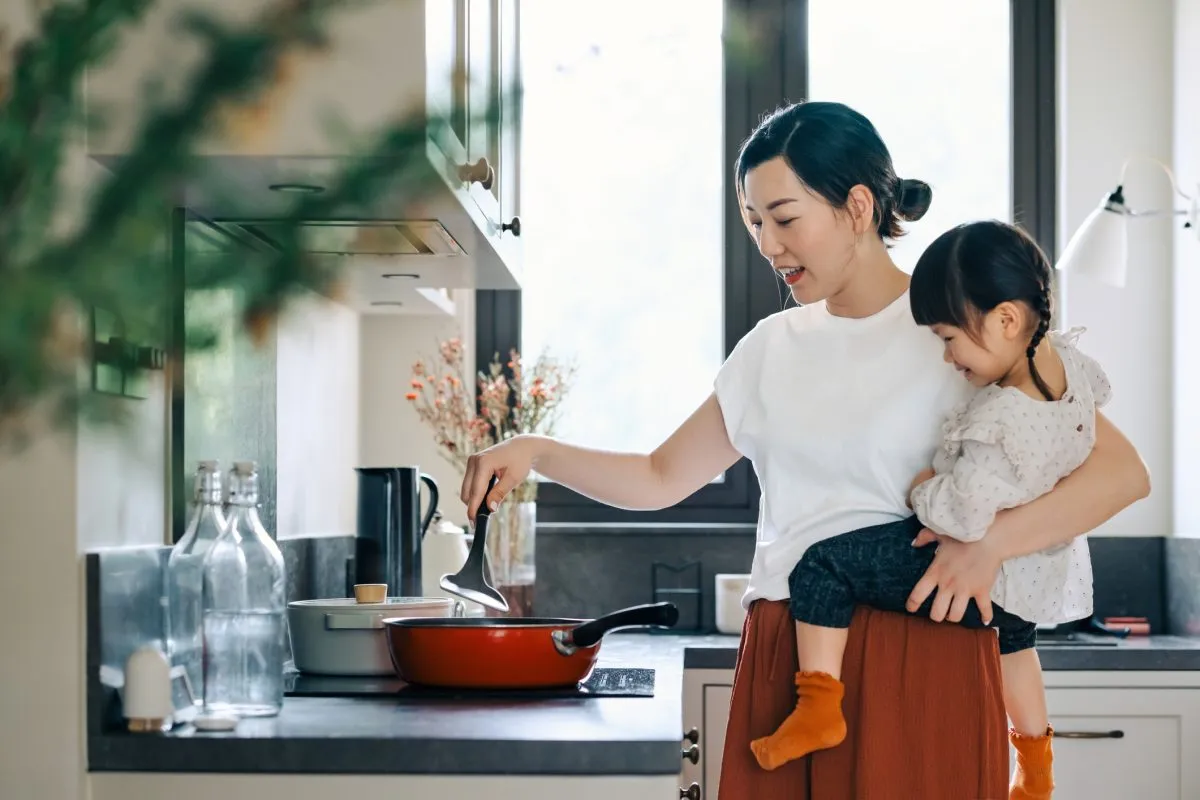 Young Asian mother carrying her daughter in arms, cooking healthy food together in the kitchen at home. Mother-daughter bonding moments. Healthy family lifestyle, bonding and togetherness