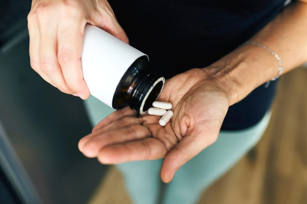 A close-up of a person dispensing white capsules from a dark bottle into their palm, illustrating medication, health care, and daily wellness management.