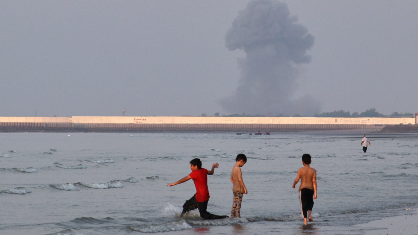 Three boys play in the shallow waters of the Strait of Hormuz, as a plume of smoke rises from an explosion in the background, off Bandar Abbas, Iran, Monday, July 13, 2026. (Razieh Poudat/ISNA via AP)