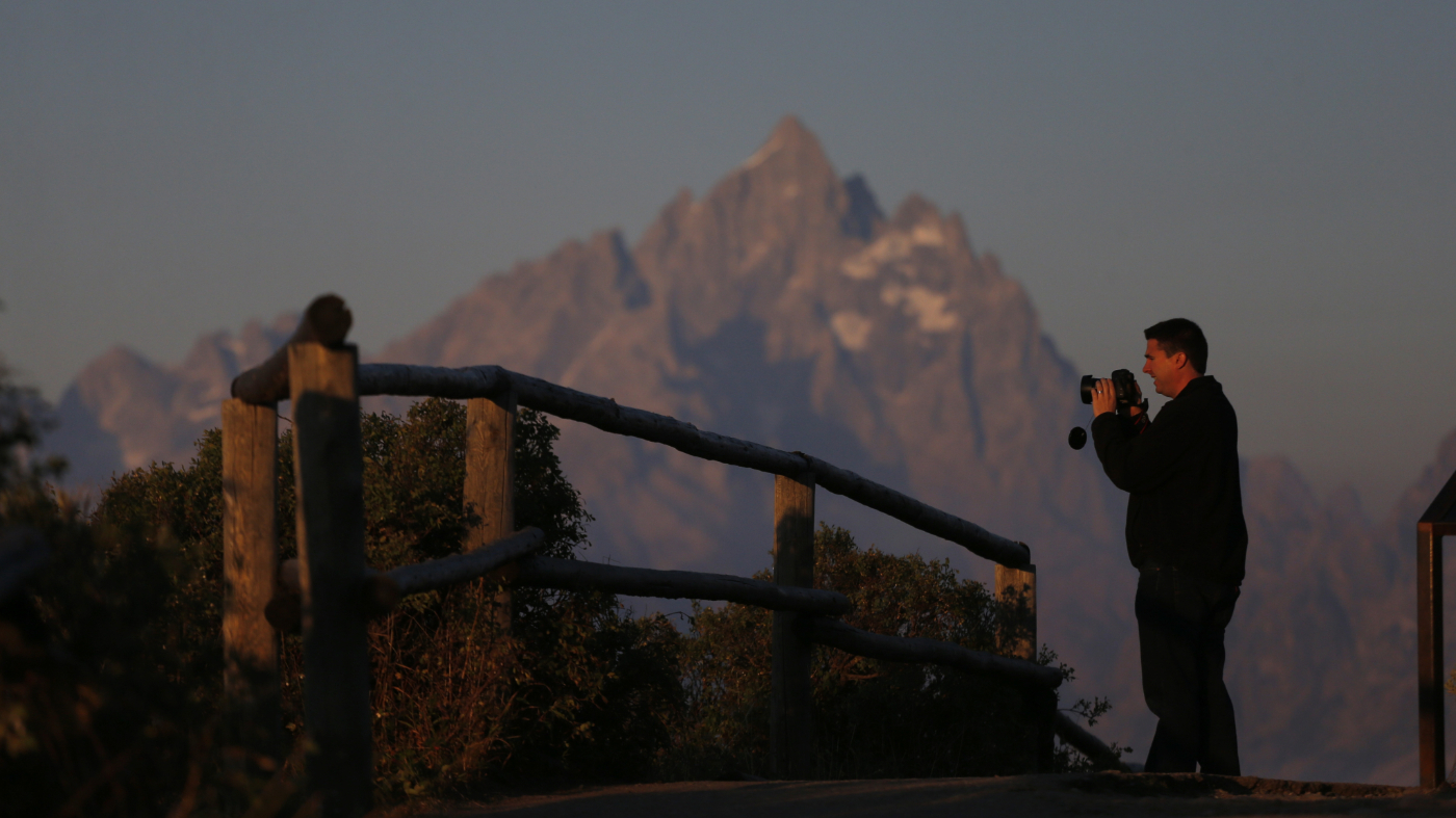 FILE – Park visitor Michael Quaide takes a picture as the sun at dawn illuminates mountain peaks, partially obscured by haze from the wildfires nearby as seen from Signal Mountain in Grand Teton National Park, Wyo., Aug 25, 2016. (AP Photo/Brennan Linsley)