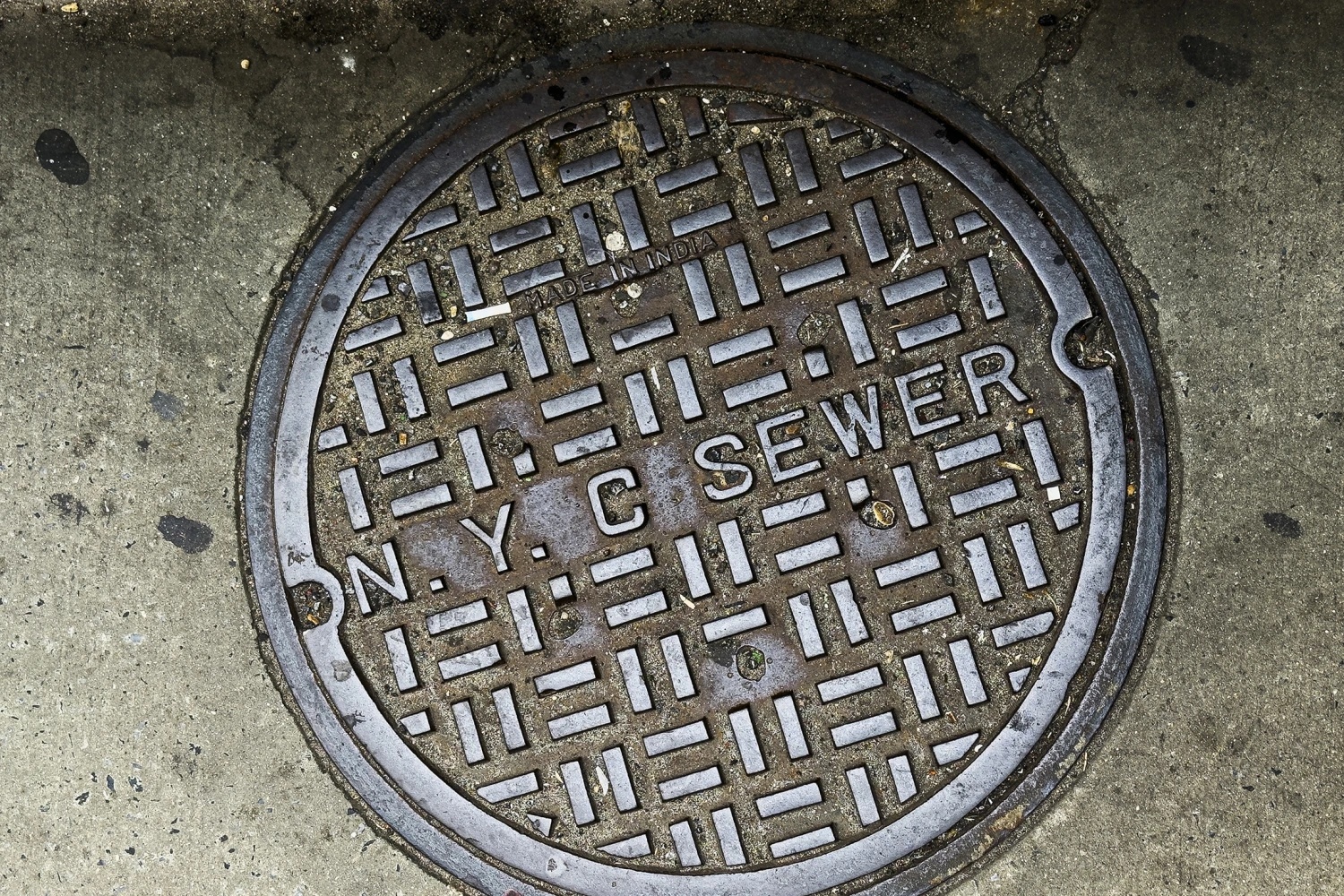 A close-up view of a classic New York City sewer cover embedded in the concrete sidewalk.