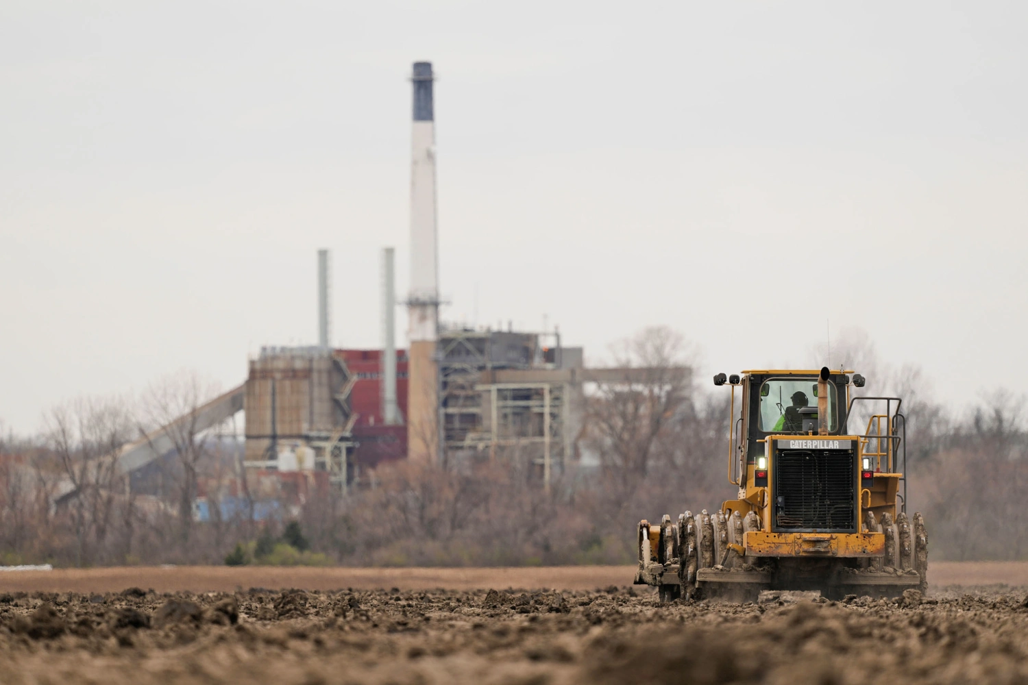 A worker prepares a plot of land for an AI data center in Independence, Mo., on March 24, 2026. A retired power plant in the background is being refurbished to provide electricity for the facility.