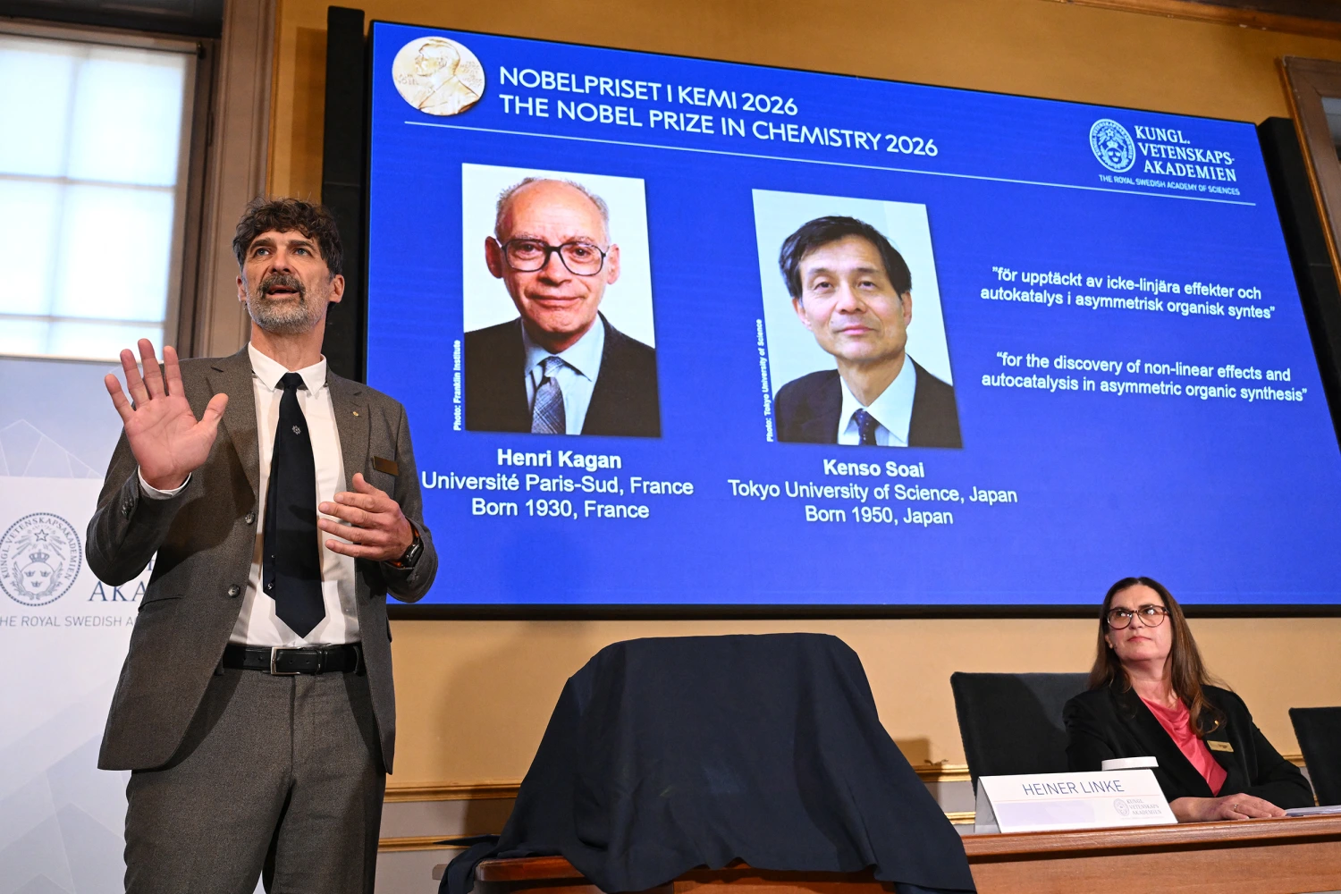 The chair of the Nobel Committee for Chemistry Heiner Linke (L) explains the field of research of Nobel Prize winners France's Henri Kagan and Japan's Kenso Soai (displayed on the screen in the background) during a press conference to announce the winners of the 2026 Nobel Prize in Chemistry at the Royal Swedish Academy of Sciences in Stockholm, Sweden, on October 7, 2026.