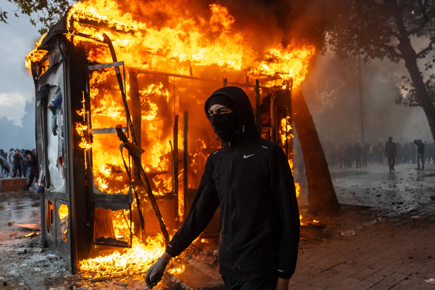 A protester walks past a burning news-stand during demonstrations on October 17, 2026 in Paris, France. The involvement of the Gilets Jaunes (Yellow Vests) with the ongoing French school blockades expands the scope of these demonstrations by linking localised student grievances to broader economic policies. Student unions originally initiated the movement to protest underfunding, deteriorating infrastructure, and acute teacher shortages across French high schools and universities. By joining the protests, the Gilets Jaunes introduce their established platform against austerity cuts, rising living costs, and surging fuel prices into the youth-led action. (Photo by Carl Court/Getty Images)