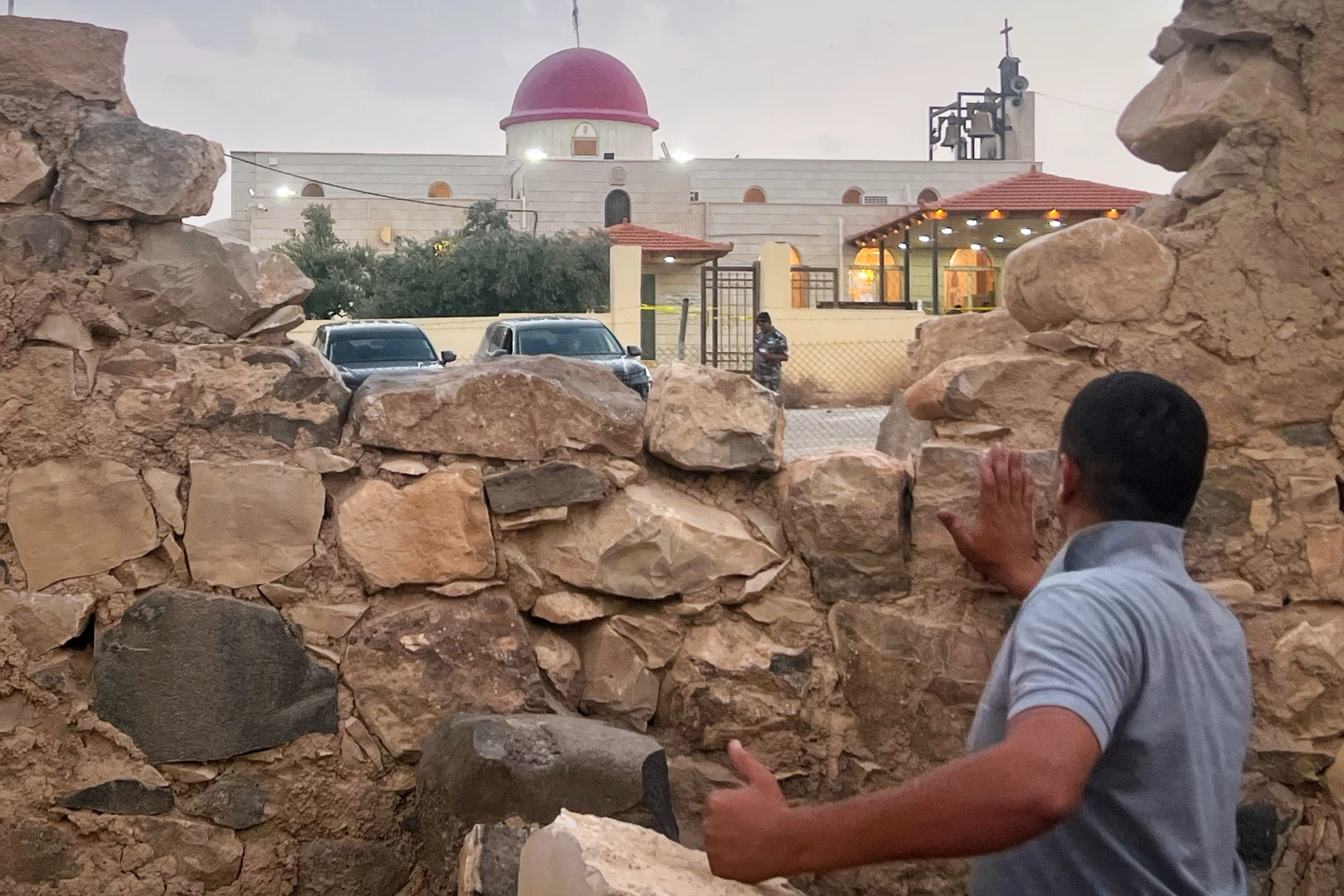 A man looks through a gap in a stone wall toward a Greek Orthodox church where gunmen opened fire during a funeral hours earlier in Hamoud village on Oct. 23, 2026.