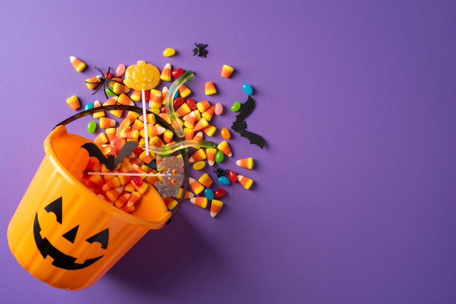 Happy Halloween scene with a jack-o'-lantern bucket spilling candy corn, lollipops, gummy worms and paper bats on a vibrant purple background conveying festive, playful seasonal fun
