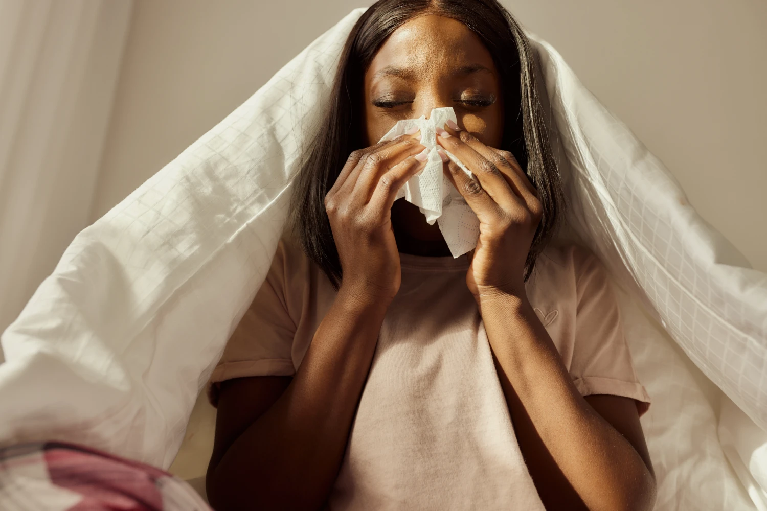 Ill African American woman blowing nose while relaxing in bedroom during morning time.
