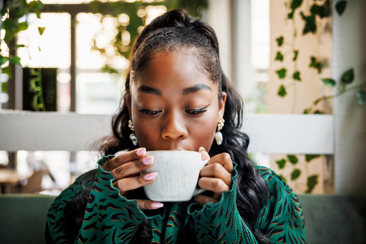 A young woman indulges in the aroma of her coffee, closing her eyes to savor the moment in a leafy cafe setting.