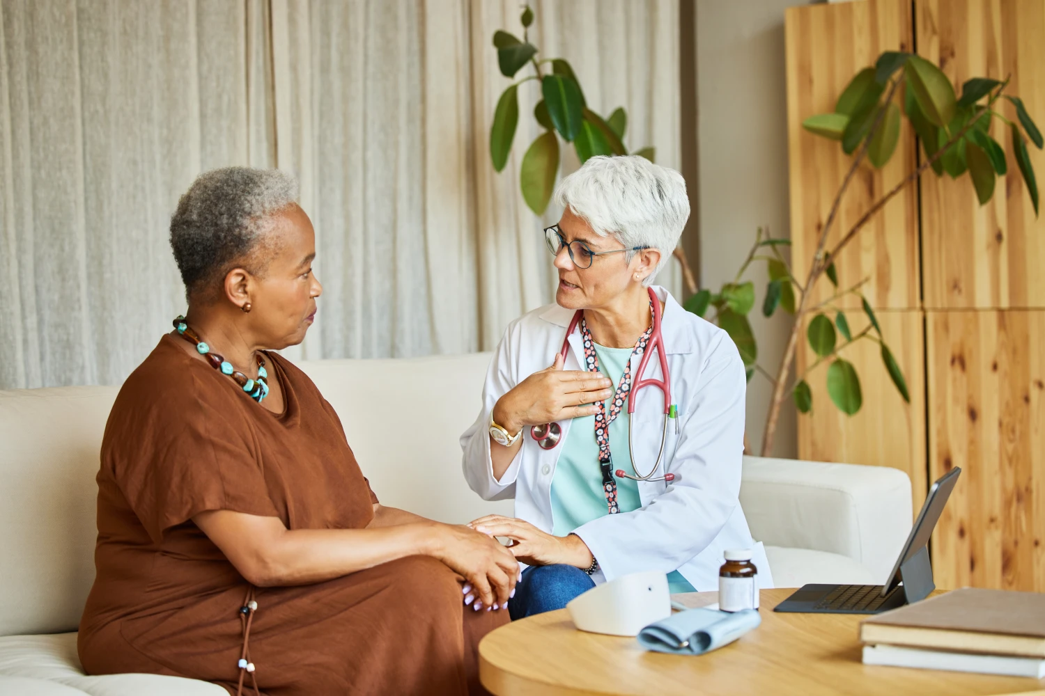 Mature doctor explaining senior woman. Female healthcare professional is visiting patient at apartment. They are sitting on sofa.