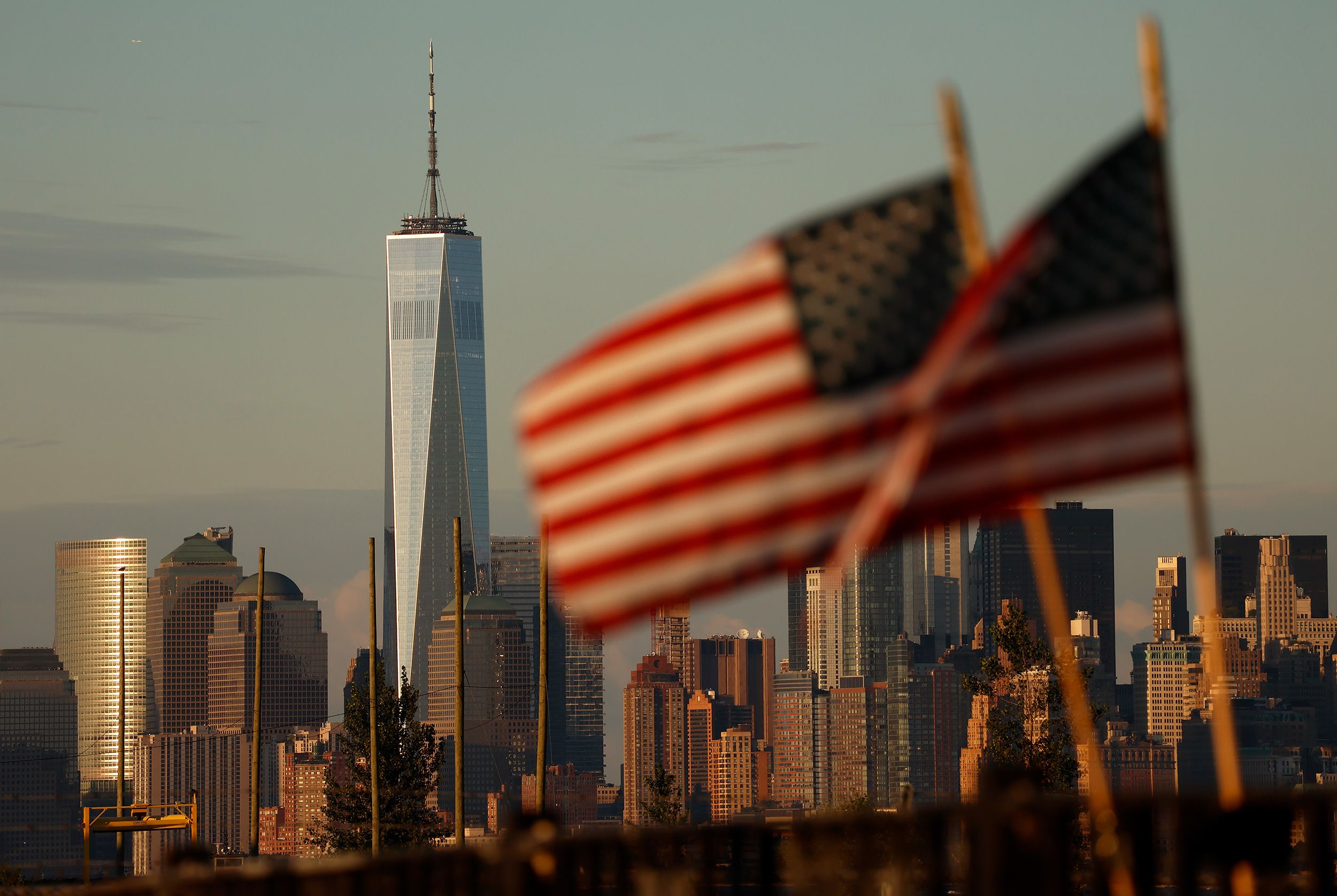BAYONNE, NJ - SEPTEMBER 1: The sun sets on the skyline of lower Manhattan and One World Trade Center in New York City as American flags fly on a fence at the Tear Drop 9/11 Memorial on September 1, 2025, in Bayonne, New Jersey.  (Photo by Gary Hershorn/Getty Images)