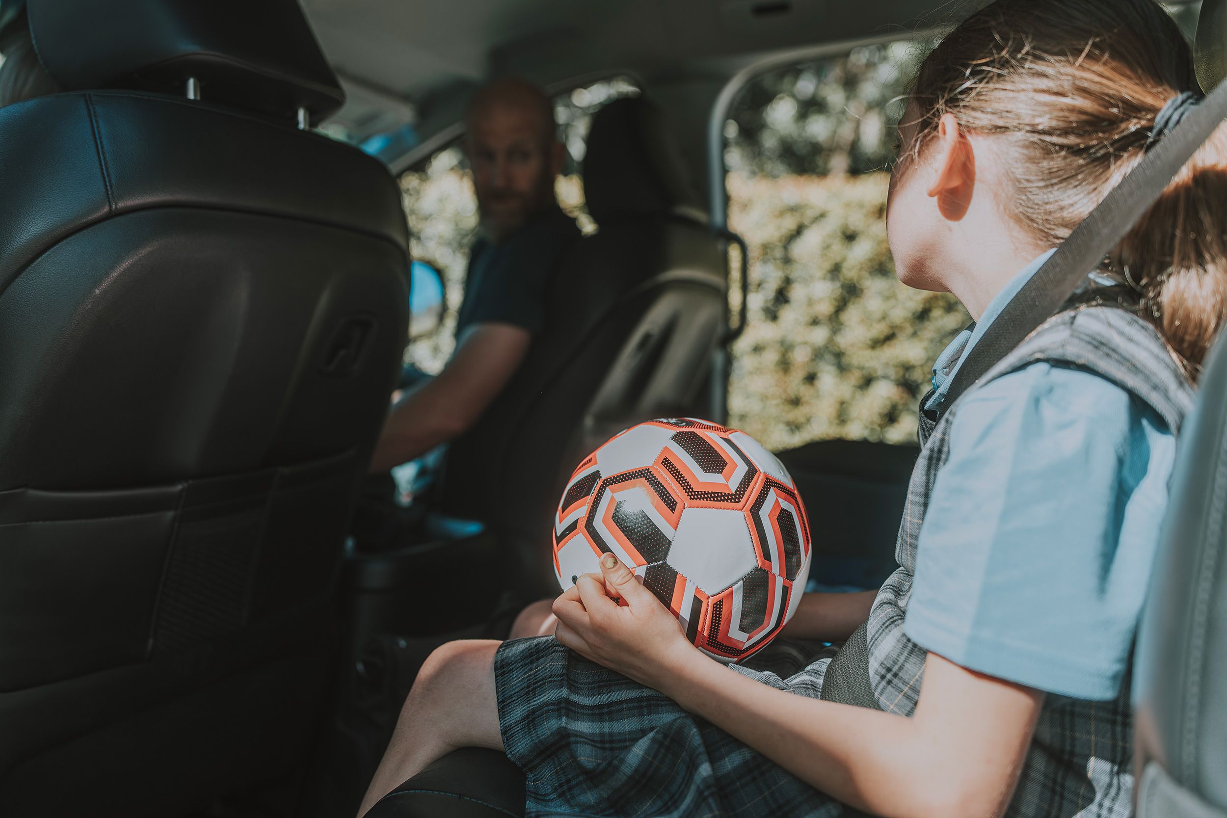 white schoolgirl in a plaid pinafore and blue uniform holds a soccer ball on her lap in the front seat as her father prepares to drive, ready for after-school sport. A candid moment linking family routine and children's sport.