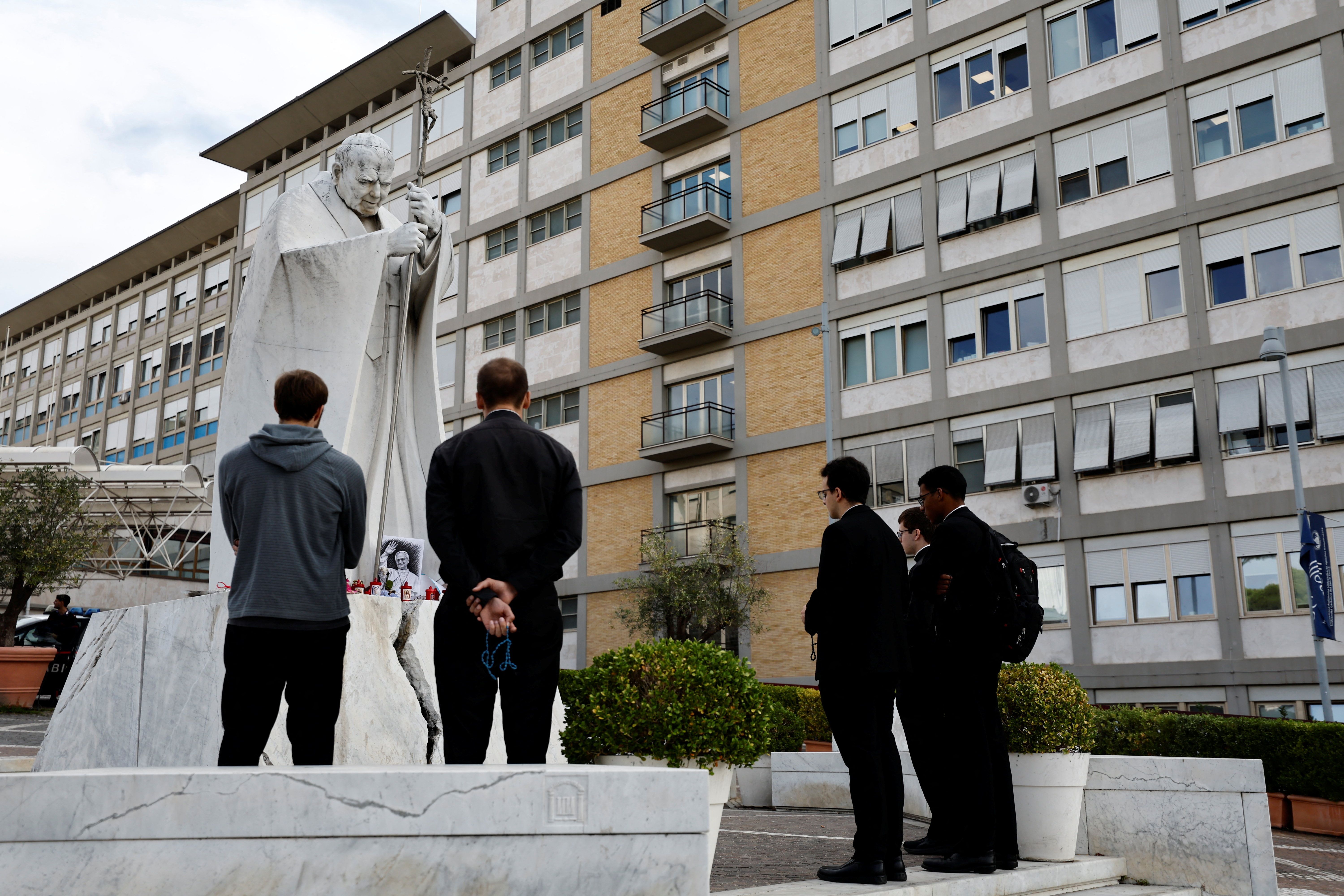 Priests and people pray the Rosary in front of the statue of the late Pope John Paul II outside Gemelli Hospital.